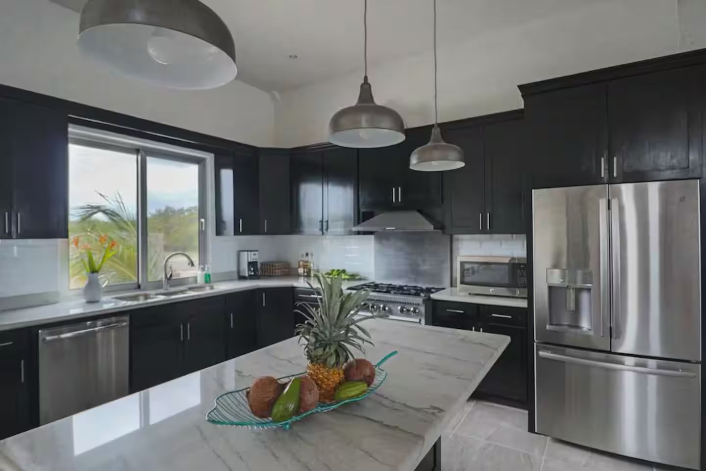 A modern kitchen with dark cabinets, stainless steel appliances, and a marble island with fruits.