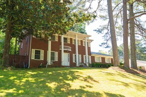 A two-story brick house with a front porch, a lawn, and trees. - Elegant Home with Spacious Bedrooms & Lush Yard (East Point)