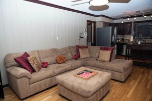 A living room with a sectional sofa, a coffee table, and a kitchen area with appliances. - Elegant Home with Spacious Bedrooms & Lush Yard (East Point)