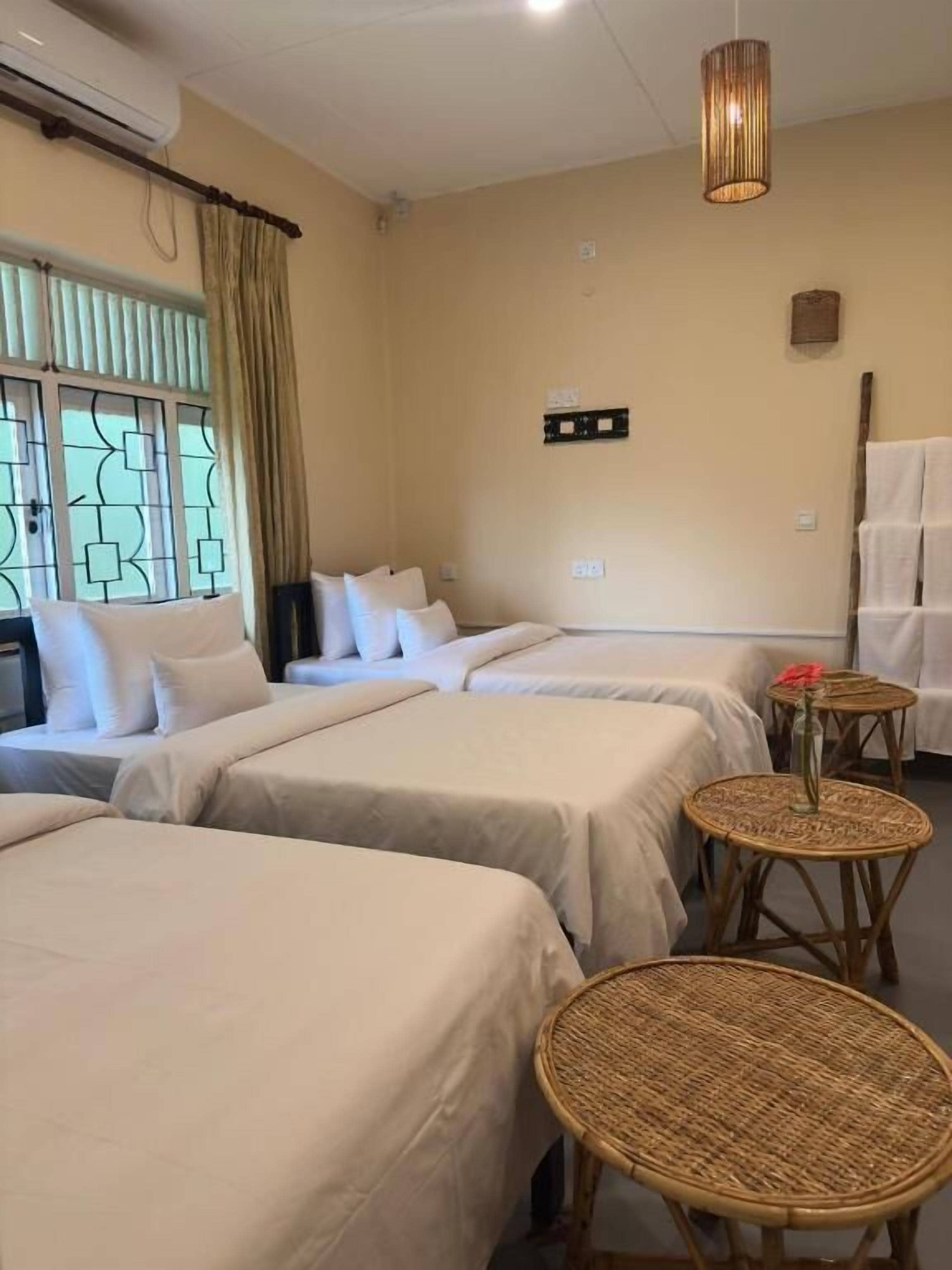 A hotel room with four beds, a large window with curtains, a wall-mounted air conditioner, a hanging light fixture, and a small table with a vase.