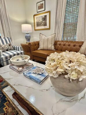 Una sala de estar bien amoblada con una mesa de centro de mármol, un jarrón grande con flores blancas, un sofá de cuero marrón, sillones a rayas y una lámpara.