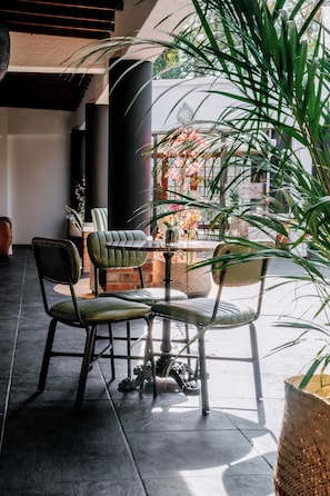 Área de estar com cadeiras e uma mesa, cercada por plantas em vasos e um painel decorativo.