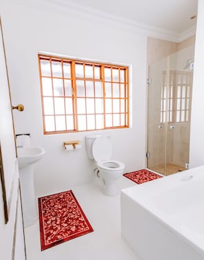 Ein Badezimmer mit Toilette, Waschbecken und Badewanne. Es gibt ein Fenster mit Holzrahmen sowie einen Duschbereich mit Glastüren.