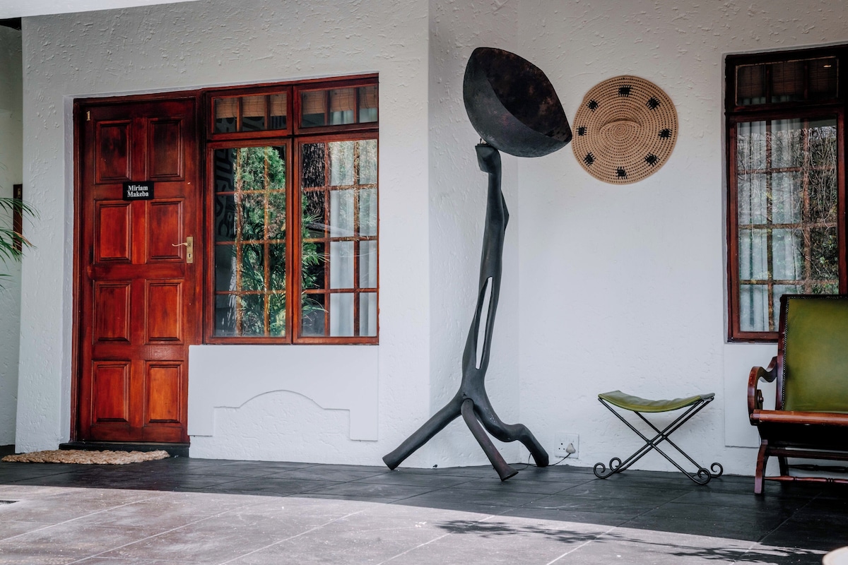 Eine traditionelle Holztür mit Namensschild, eine große Metallskulptur, ein gewebter Hut und ein grüner Sitzhocker.