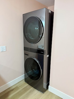 A compact, modern, two-door washer and dryer unit with a stainless steel finish, positioned in a corner against a light-colored wall.