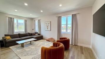 A modern living room with a sectional sofa, a coffee table, two ottomans, and large windows with curtains.