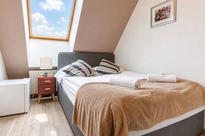 A bedroom with a bed, bedside table, lamp, and a window with a view of the sky.