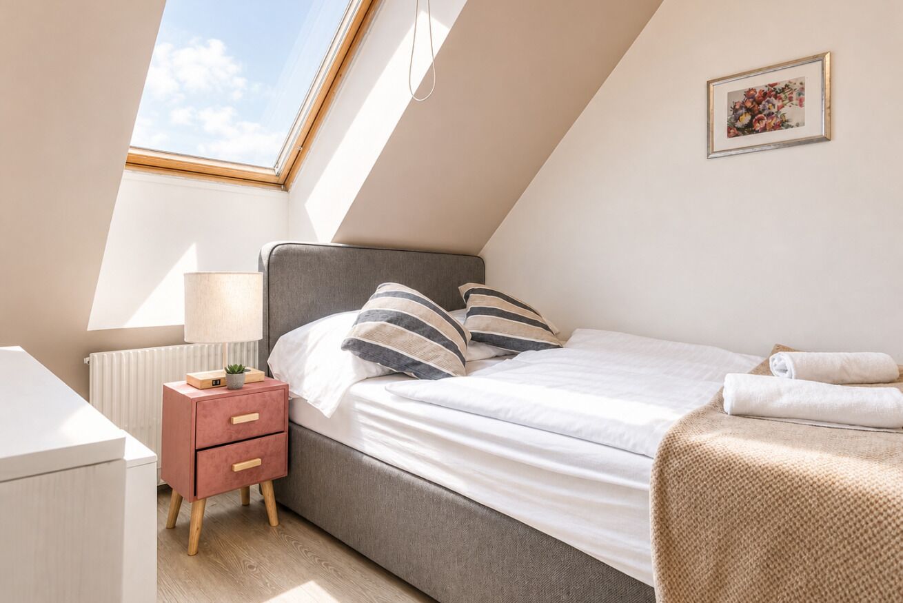 Une chambre avec un puits de lumière, un lit avec des coussins à rayures, une table de chevet avec une lampe et un radiateur.