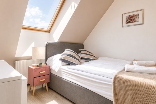 A bedroom with a skylight, a bed with striped pillows, a bedside table with a lamp, and a radiator.