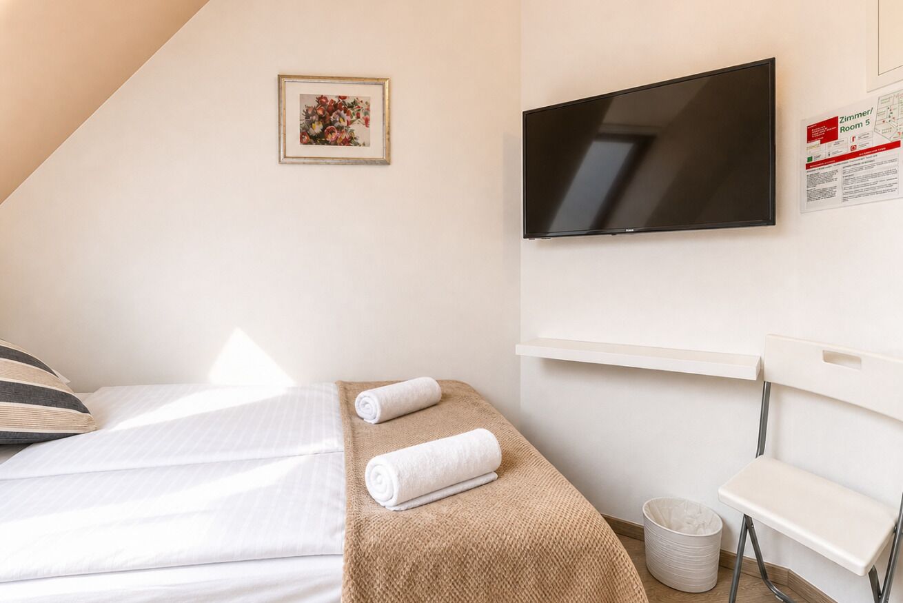 Ett hotellrum med en säng, en tv som är monterad på väggen, ett litet bord, en stol och en papperskorg.
