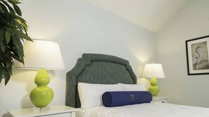 A hotel room with a bed, two bedside tables, two lamps, a pillow, a framed picture on the wall, and a plant.
