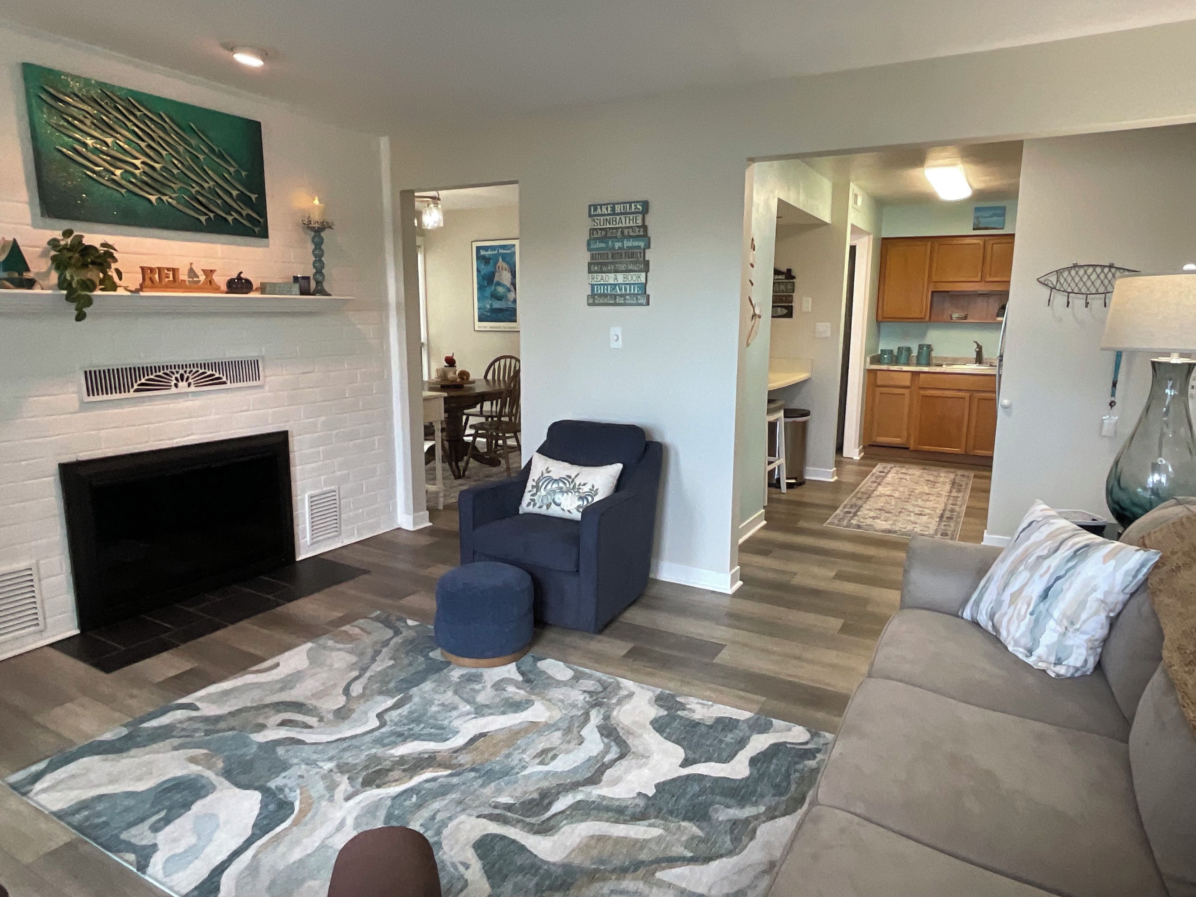 A living room with a fireplace, a blue armchair, a patterned rug, and a kitchen in the background.