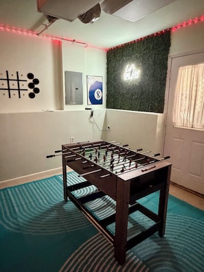 Quarto com mesa de futebol, jogo de tabuleiro na parede e parede verde com letreiro neon.