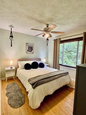 Un dormitorio con cama, mesita de noche, ventilador de techo, ventana con cortinas e imagen colgada en la pared.