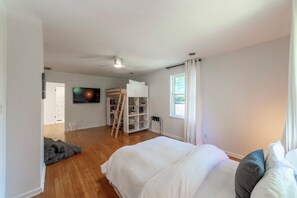 Un dormitorio con cama, televisión montada en la pared, estantería y ventana con cortinas.