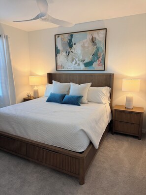 A bedroom with a large bed, two bedside tables, a ceiling fan, and a painting on the wall.