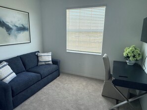 A room with a dark blue sofa, a desk, a window with blinds, and a framed picture on the wall.
