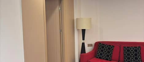 A hotel room with a wooden door, a red sofa with patterned cushions, a black table with a tissue box and a small item, and a floor lamp.