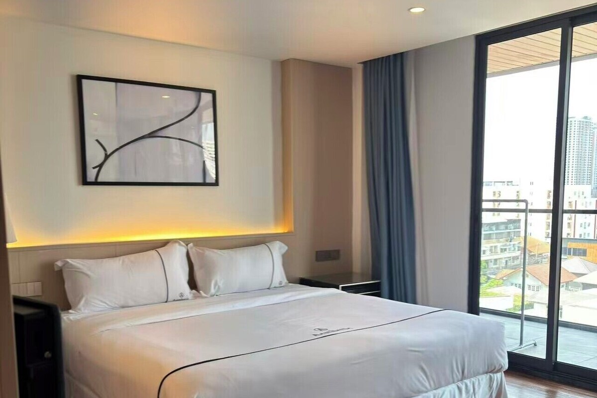 A hotel room with a large bed, a nightstand, a wall-mounted abstract art piece, and a view of the city through a large window.