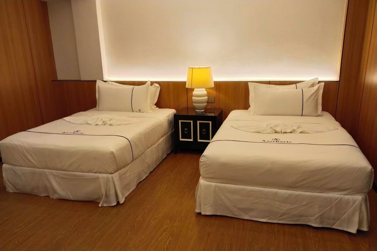 A hotel room with two beds, a nightstand with a lamp, and wooden paneling on the walls.
