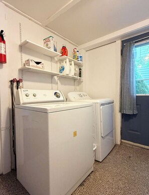 A laundry room with a front-loading washer and dryer, shelves with cleaning supplies, and a fire extinguisher on the wall. - Webster’s Place — A charming Bar Harbor home (Bar Harbor)