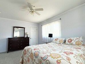 A bedroom with a bed, dresser, mirror, ceiling fan, window with curtains, and a door. - Webster’s Place — A charming Bar Harbor home (Bar Harbor)