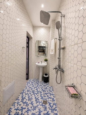 Casa de banho com paredes em padrão hexagonal, lavatório com coluna, espelho, chuveiro com chuveiro manual e chão com azulejos azuis e brancos.