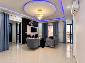 A modern living room with a flat-screen TV, two sofas, a ceiling light fixture, and a large window with curtains.