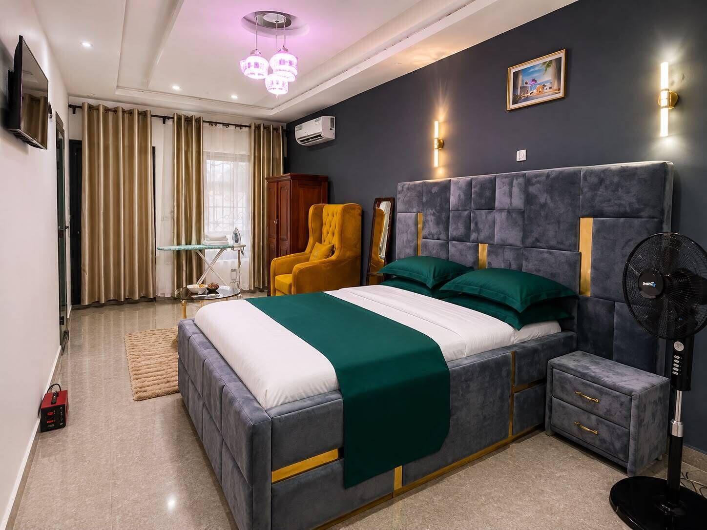 Ein modernes Schlafzimmer mit einem großen Bett, Nachttischen, einem Sessel, einem Standventilator und einem Bild an der Wand.
