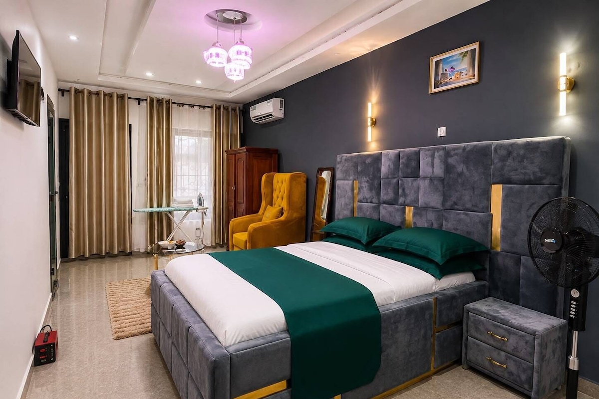 Quarto moderno com uma cama grande, mesas de cabeceira, uma cadeira, um ventilador de pé e um quadro na parede.