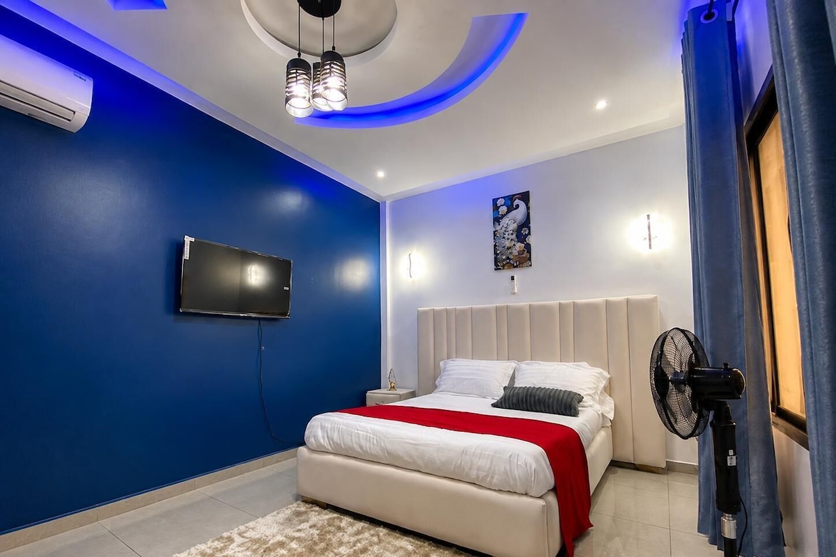 Quarto moderno com cama, televisão de tela plana, ventilador de teto e um detalhe decorativo na parede.