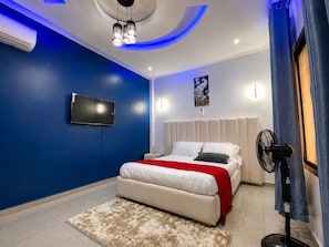 Quarto moderno com cama, televisão de tela plana, ventilador de teto e um detalhe decorativo na parede.