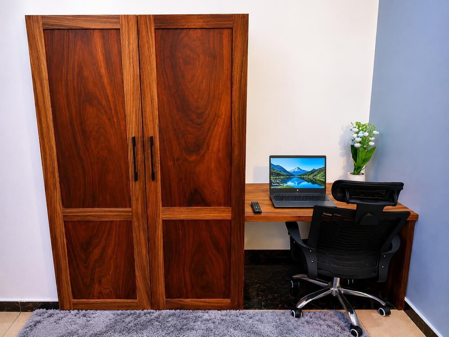 A room with a wooden wardrobe, a desk with a laptop, a chair, and a small potted plant.