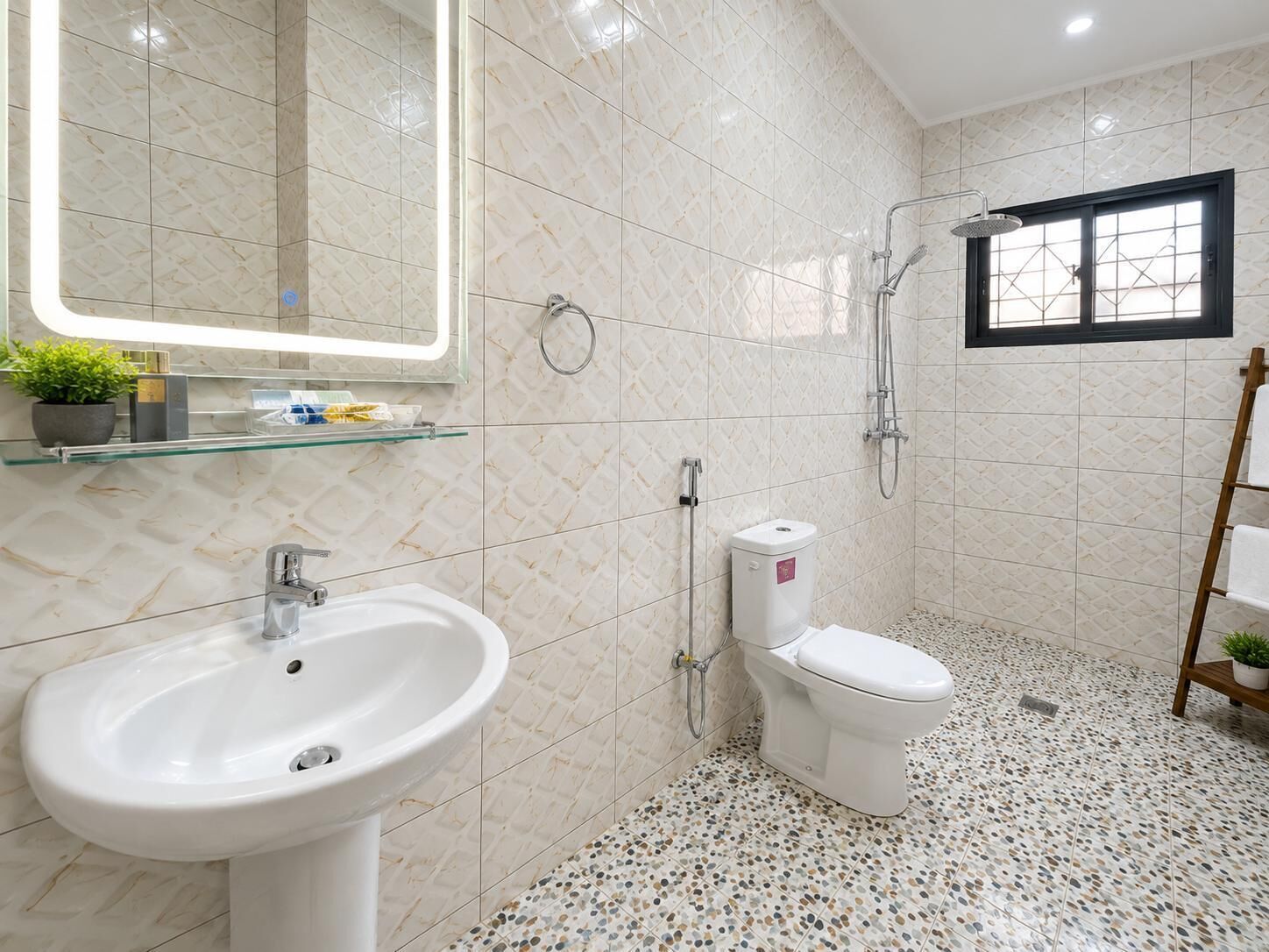 Baño con lavamanos blanco, inodoro y ducha. Se observa una repisa de cristal con artículos de aseo y una planta en maceta. El suelo presenta un diseño de baldosas con textura.