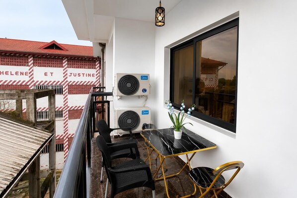 A balcony with two air conditioning units, a table with chairs, and a window with a view of buildings. - Nova vista home (Douala)