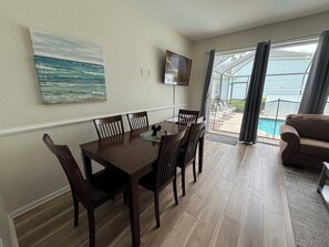 Ein Essbereich mit einem Holztisch und Stühlen, einem Meeresbild, einem Fernseher und Blick auf einen Pool durch große Fenster.
