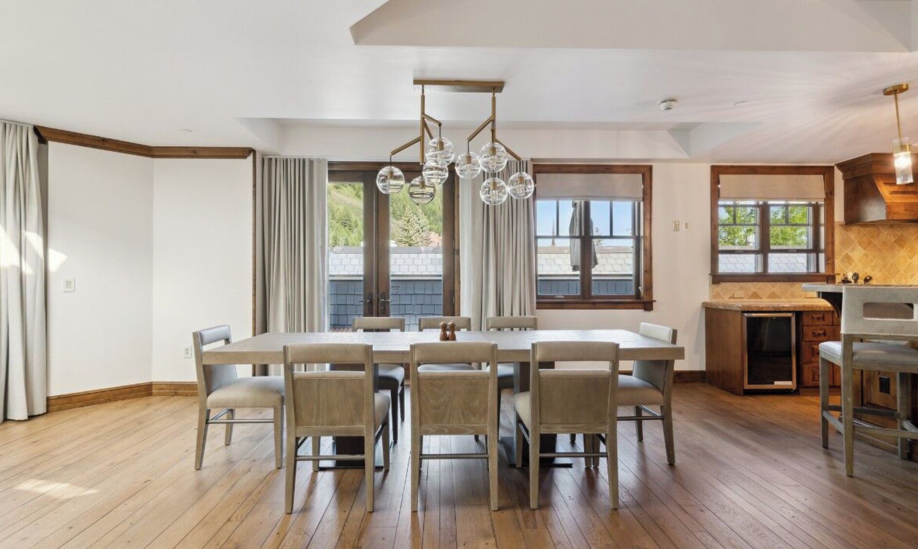 A spacious dining area with a long table and chairs, a modern chandelier, and large windows offering views of the outdoors.