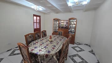 Une salle à manger avec une table ronde, des chaises en bois, une nappe à motifs floraux, un buffet aux portes vitrées et un ventilateur de plafond.