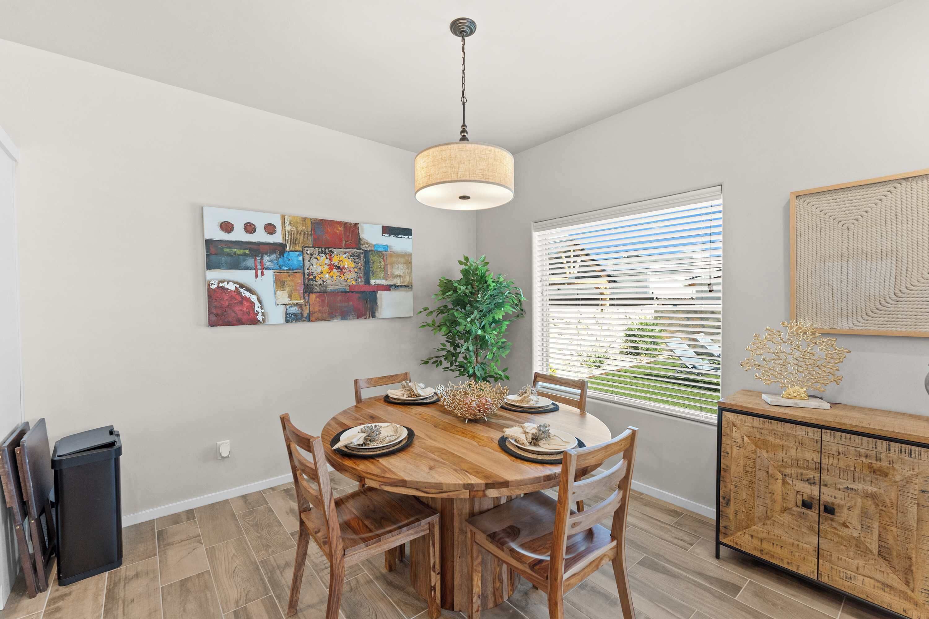 A dining area with a wooden table, chairs, a hanging light fixture, and a window with blinds.