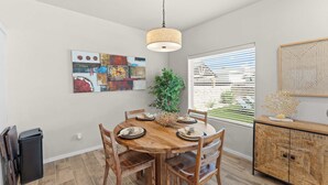 Een eetgedeelte met een houten tafel, stoelen, een hanglamp en een raam met jaloezieën.