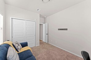 A hallway with a closed white door, a white wall with a black sign, a blue couch with a yellow throw, and a carpeted floor.