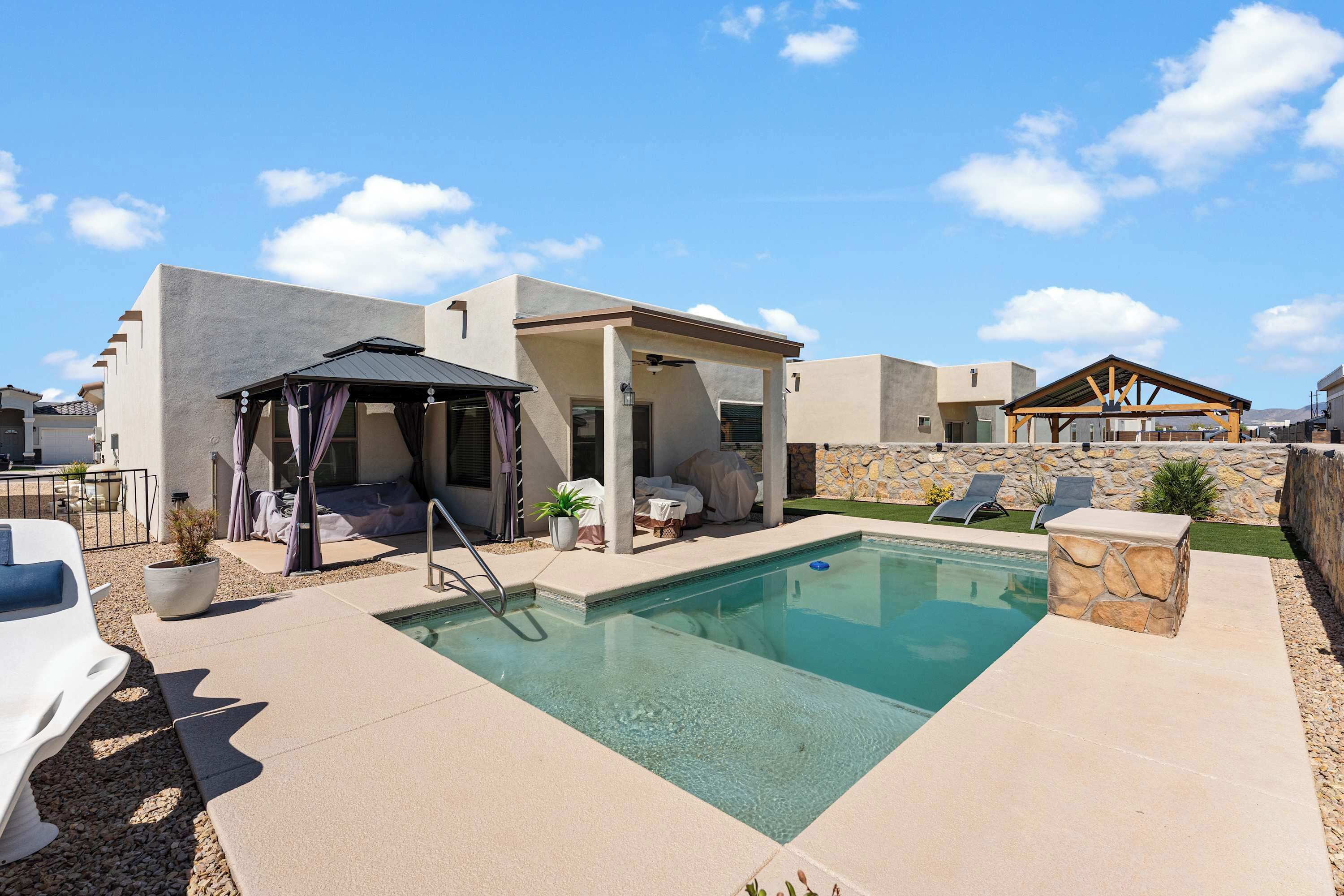 Un área de piscina con zona de estar cubierta, sillas de descanso y un muro de piedra.