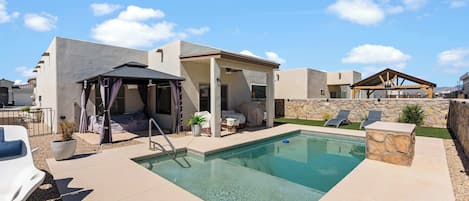 Un área de piscina con zona de estar cubierta, sillas de descanso y un muro de piedra.
