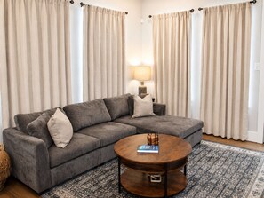 Een woonkamer met een grijze hoekbank, een ronde houten salontafel en een vloerkleed met een patroon.