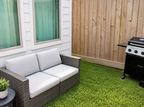 Een rieten bank met kussens, een klein rond tafeltje met een plant in pot, en een barbecue.