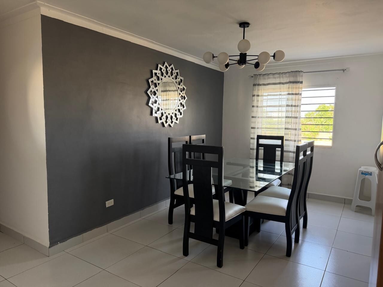 Un coin repas avec une table et des chaises en verre, un élément décoratif mural et une fenêtre avec des rideaux.