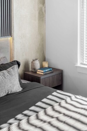 A bedroom with a bed, a bedside table with books and a candle, and a window with blinds. - The Dominic - 1BR Condo, King Bed, Pool, Gym (Orlando)