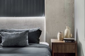Ein Schlafzimmer mit einem Bett, Kissen, einem Nachttisch mit Büchern und einer Vase sowie einem Fenster mit Jalousien.