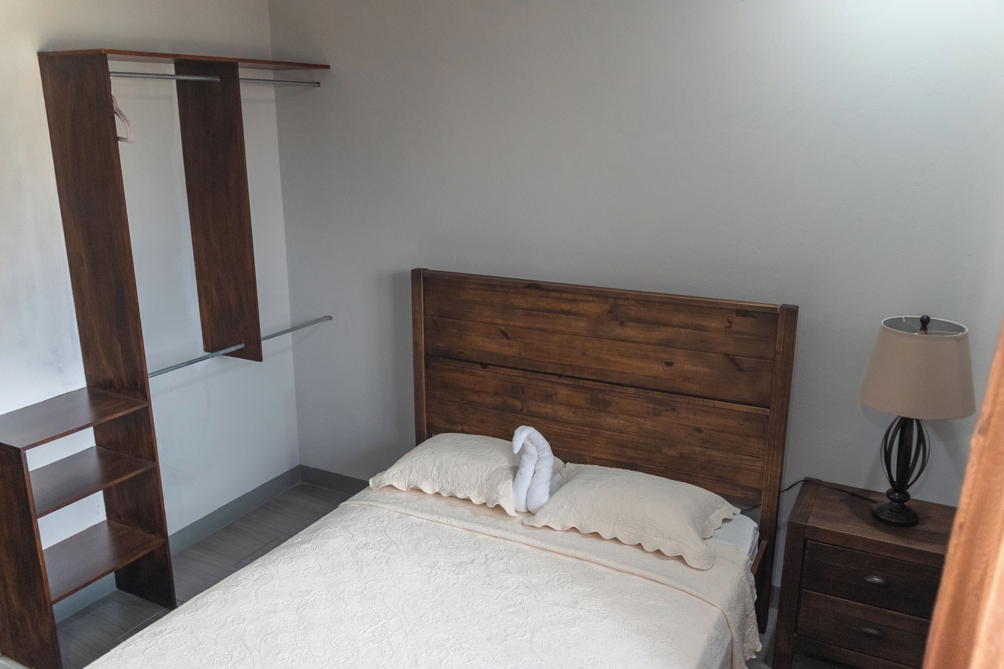 Un dormitorio con cabecera de madera, cama con cubrecama blanco, mesita de noche con lámpara y armario de madera con espejo.