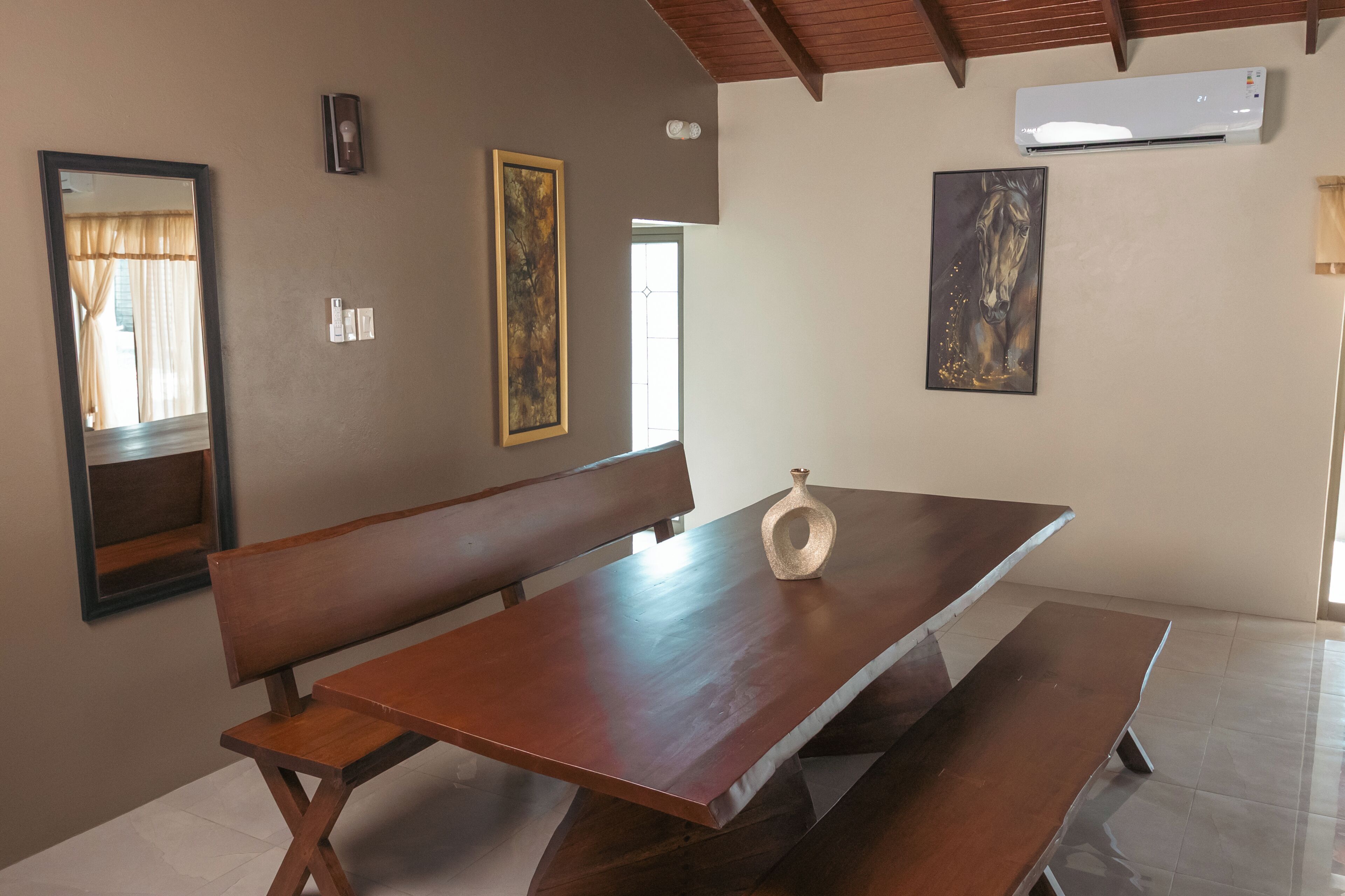 A dining area with a wooden bench, a table, and a vase. There are framed artworks on the walls and an air conditioning unit above.